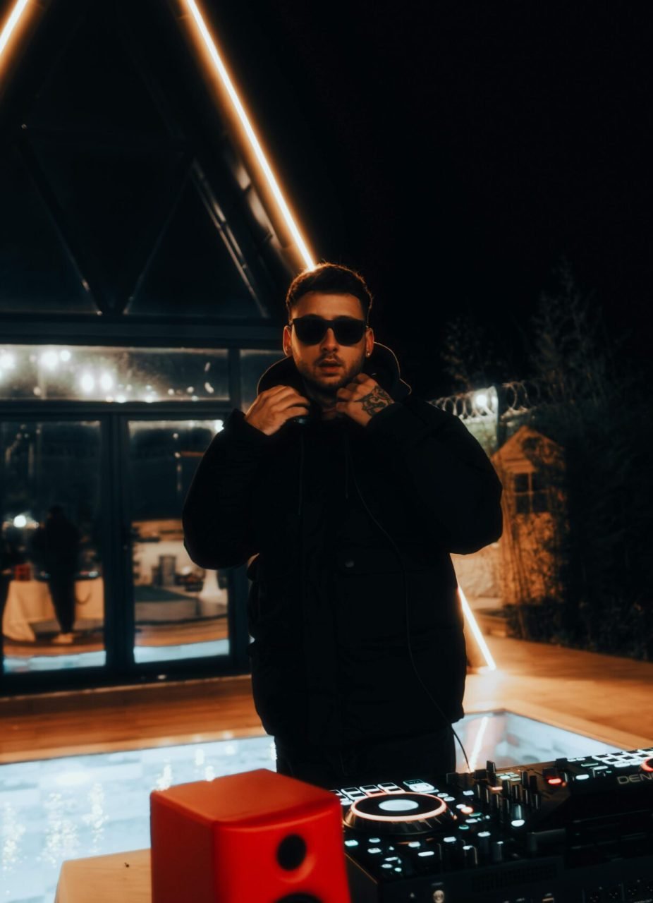 A DJ in sunglasses mixing tracks at a stylish nighttime pool party with vibrant lighting.